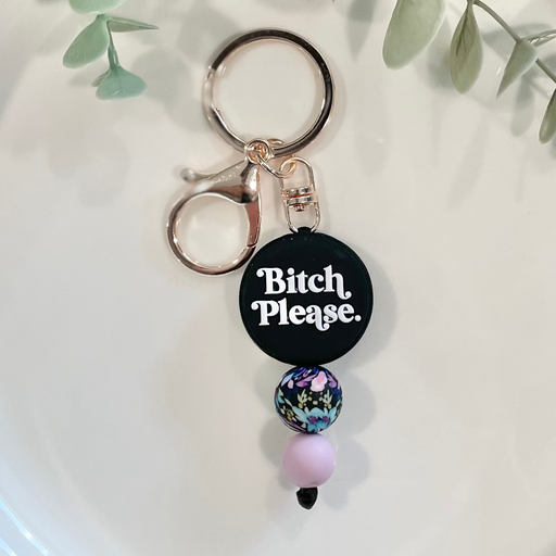 A beaded keychain with a circular charm that has the phrase 'Bitch Please' written on it, displayed on a white plate with green leafy branches in the background.