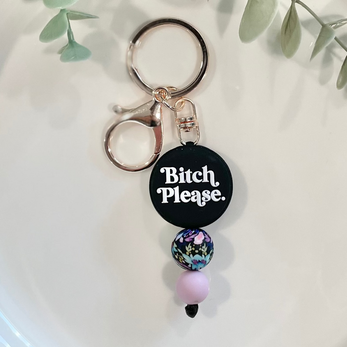 A beaded keychain with a circular charm that has the phrase 'Bitch Please' written on it, displayed on a white plate with green leafy branches in the background.