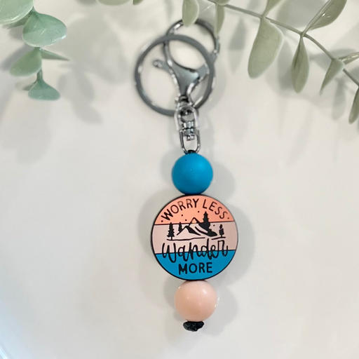 A beaded keychain with a circular charm featuring text, placed on a white plate with green leafy branches in the background.