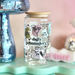 Glass jar with decorative text and illustrations on a light blue stand with a pink background